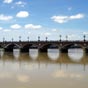 Nous allons franchir le pont de Pierre qui conduit au centre de Bordeaux. L'absence de pont sur la Garonne a longtemps été considérée par les Bordelais comme une défense naturelle face à l'ennemi. Depuis la réalisation du premier ouvrage en 1821, le pont de Pierre, le désenclavement de la ville a été une priorité vitale. Après la passerelle Eiffel en 1860, il faudra néanmoins attendre plus d'un siècle pour construire un nouveau pont. Il relie la rive gauche au quartier de la Bastide. Premier pont sur la Garonne, il fut construit sur ordre de Napoléon Ier entre 1810 et 1822, d'après les plans de Claude Deschamps. Il mesure 486 m de longueur et 19 m de largeur. Les premières assises de la maçonnerie des piles ont été mises en place grâce à des caissons compartimentés de 23 m de longueur, de 7,40 m de largeur et de 6 m de hauteur. Durant douze ans les bâtisseurs durent faire face à de nombreux problèmes en raison du courant très fort à cet endroit-là. C'est grâce à une cloche à plongée empruntée aux Britanniques que les piliers du pont purent être stabilisés. Il compte dix-sept arches (nombre de lettres dans le nom de Napoléon Bonaparte). Sur les côtés, chaque pile de briques est rehaussée d'un médaillon blanc en l'honneur de l'empereur. Il porte aussi les petites armoiries de la ville (les trois croissants entrelacés). Ce pont fut le seul jusqu'à la construction du pont Saint-Jean en 1965.